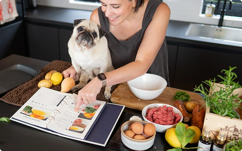 Woman with her pug reading the LOONAWELL Cookbook in a kitchen while preparing homemade dog food with fresh meat, eggs and vegetables.