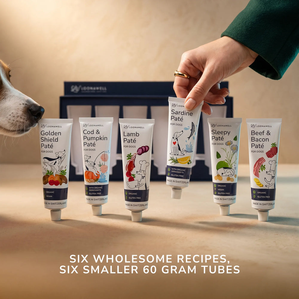 Six tubes of Loonawell Pates, all different recipes, are lined up on a wooden surface, with a hand holding up the Sardine Pâté tube and a dog's face visible on the left sniffing one of the pates. The background is a warm beige wall.