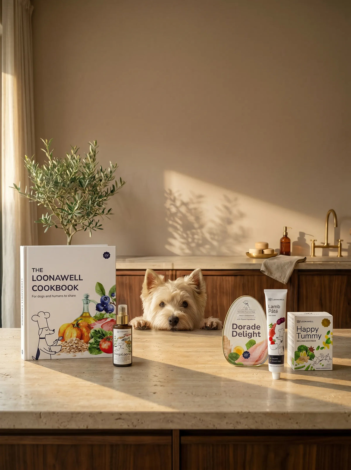A small white Westie dog peers over a kitchen counter where several packaged products are arranged, including a cookbook titled 'The Loonawell Cookbook,' a small bottle, a round item labeled 'Dorade Delight,' a tube, and a box labeled 'Happy Tummy.' The scene is styled in a warm, sunlit kitchen with a plant, countertop shadows, and cabinetry in the background.