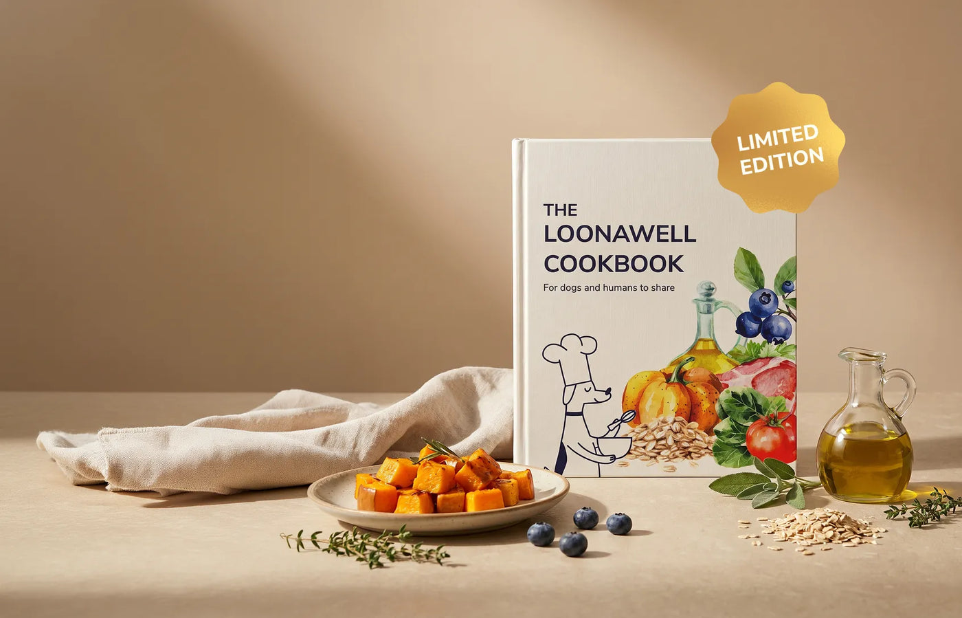 The Loonawell Cookbook standing upright next to a plate of roasted sweet potatoes, fresh blueberries, herbs, oats, and olive oil. A gold badge on the book reads "Limited Edition," and the cover text says "The Loonawell Cookbook: For dogs and humans to share."