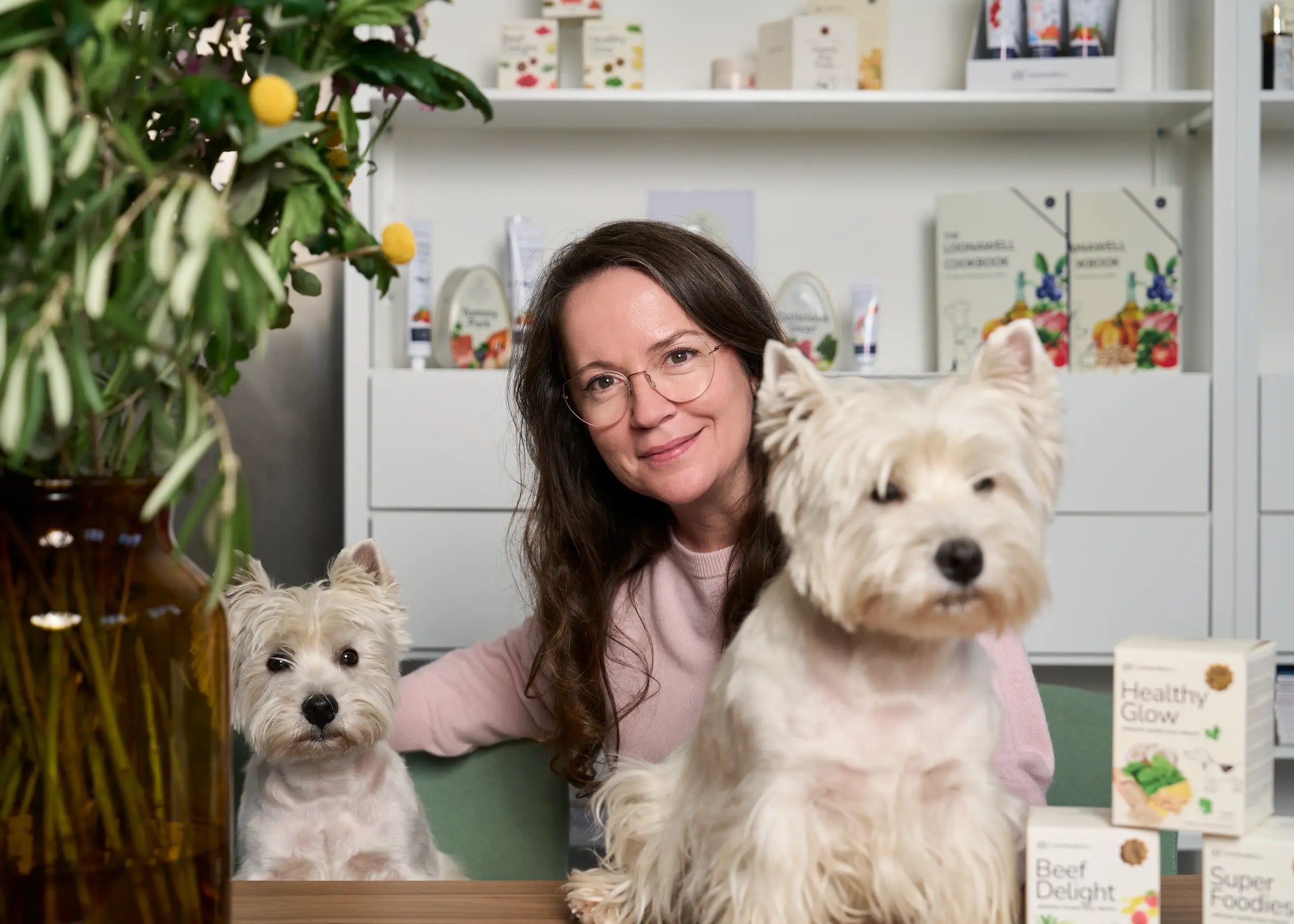 Maria, the founder of Loonawell, smiling warmly at a table alongside her two West Highland White Terriers. Several Loonawell dog treats and supplements are displayed on the table and on the shelves behind her.